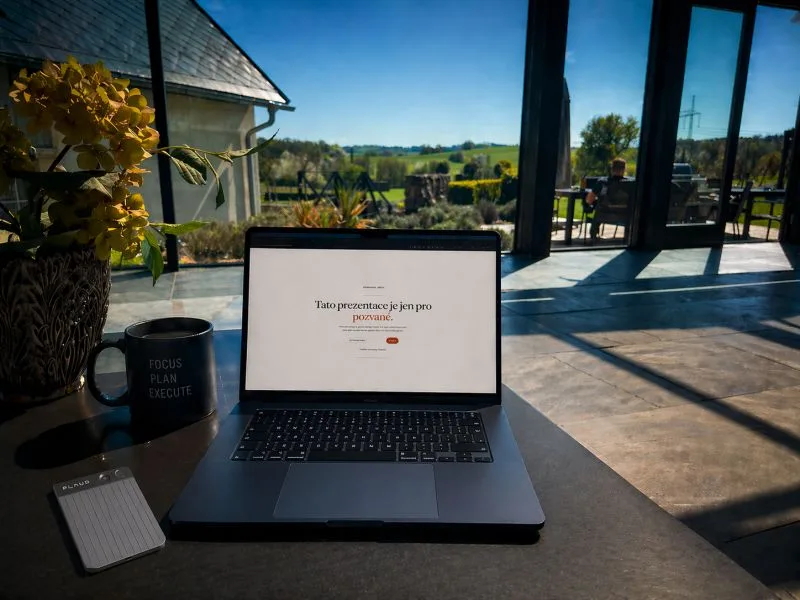Notebook a hrnek na stole u prosklené stěny, vedle váza se žlutými květy, za sklem zahrada a krajina